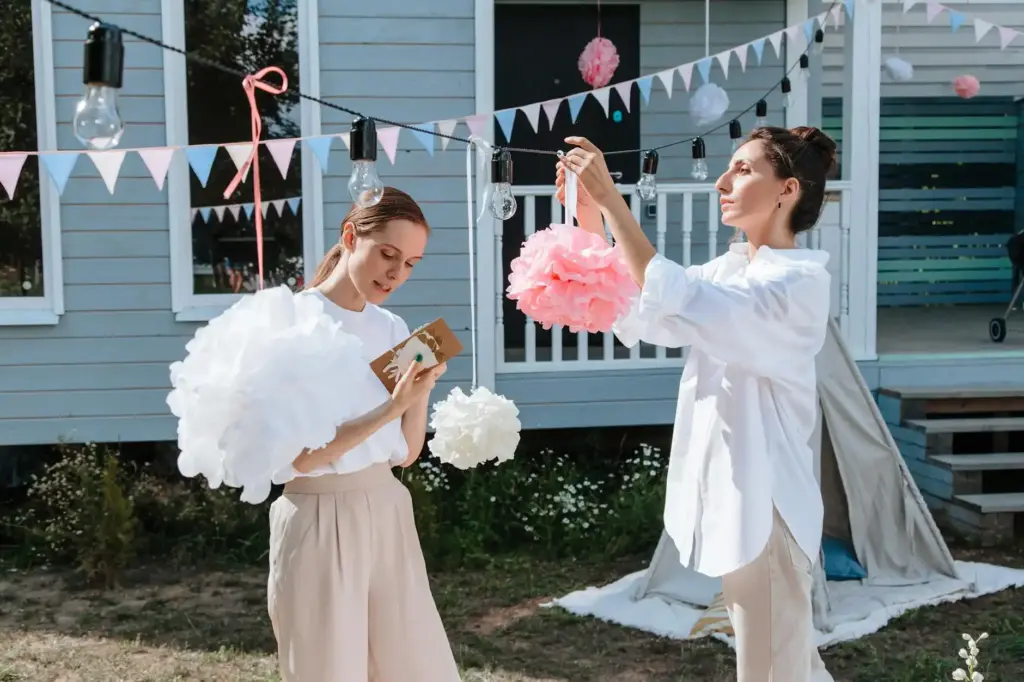 Two women in a backyard preparing party decorations with pom-poms and lightbulbs.