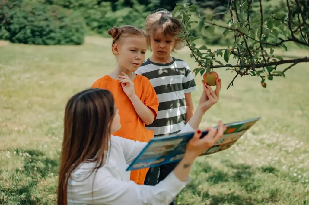 Young children learning outdoors with a teacher, exploring nature and reading a book.