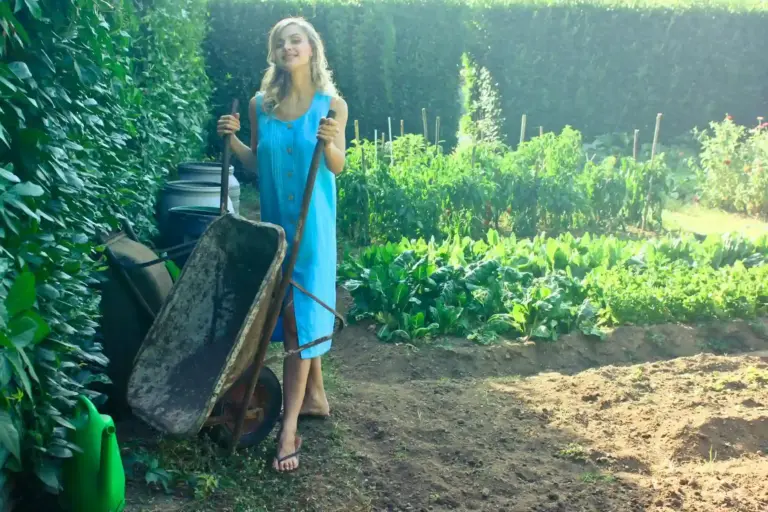 A woman enjoys gardening in her lush home garden beside a wheelbarrow and crops.