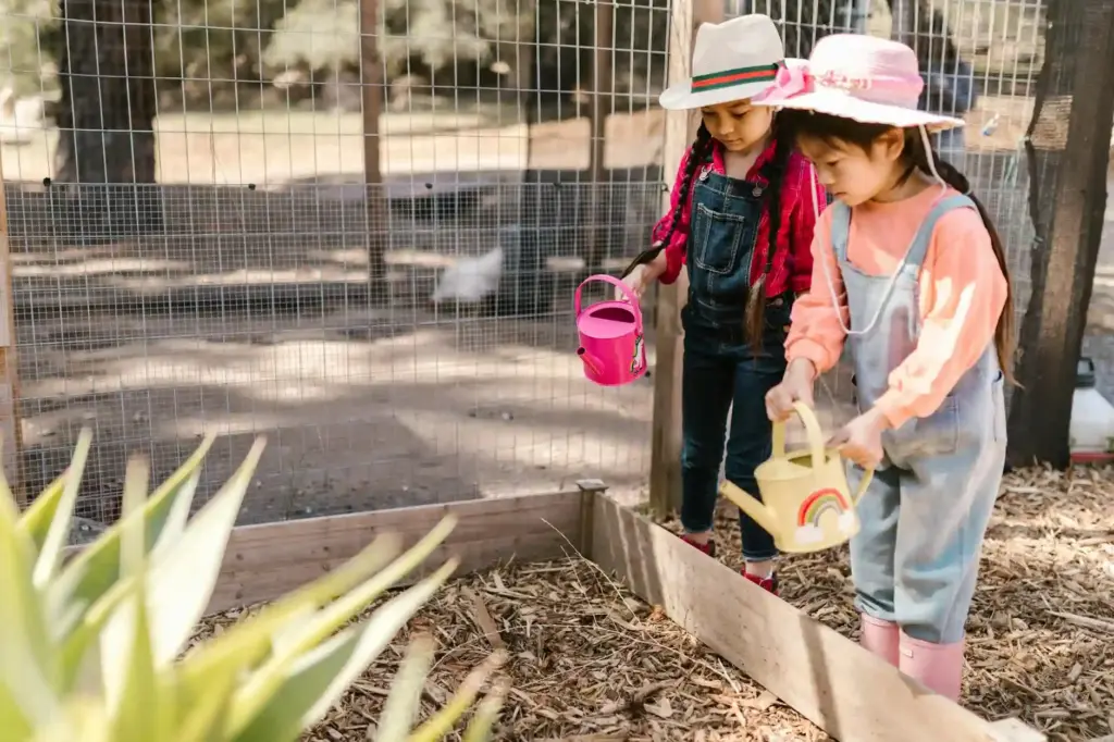 Two young girls gardening outdoors with watering cans and sun hats, fostering nature appreciation.