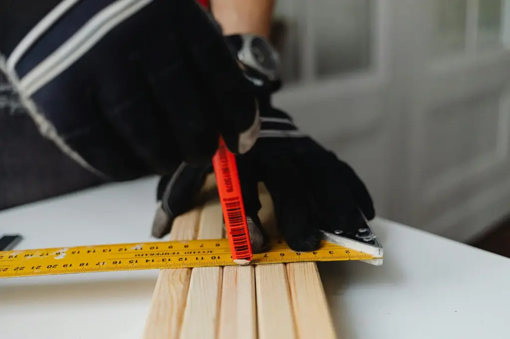 Close-up of a gloved carpenter measuring wood indoors using a ruler and pencil.