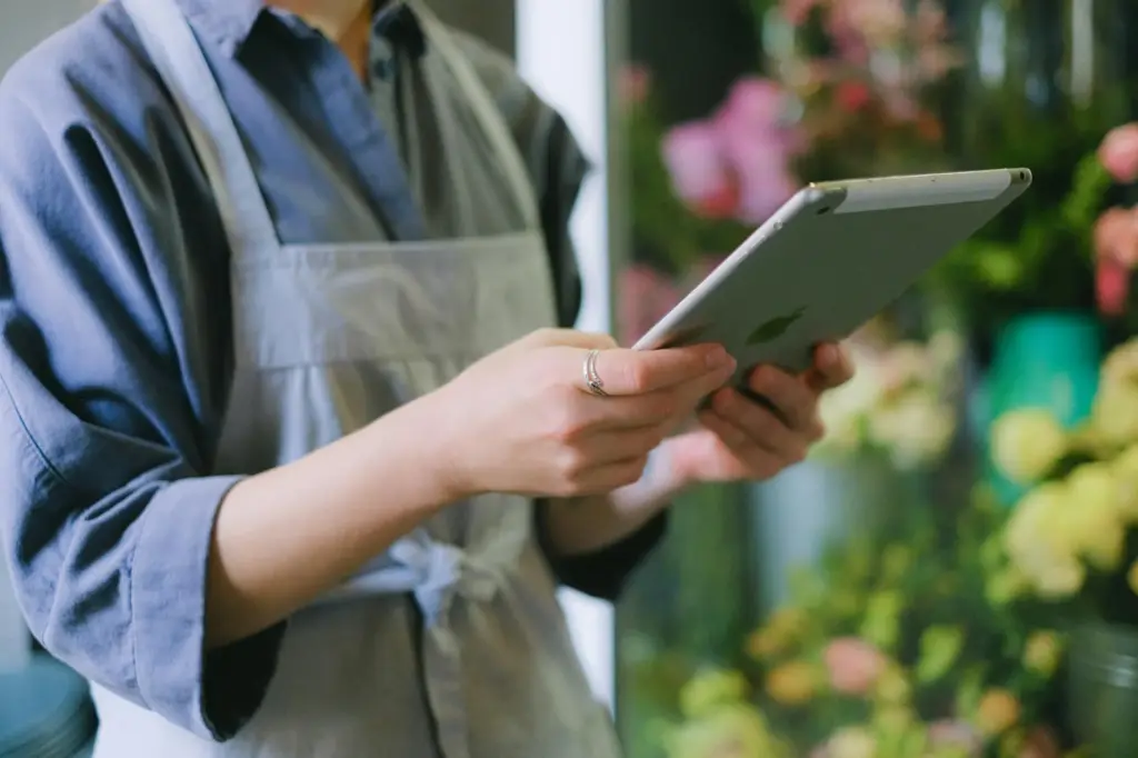 Florist in apron using digital tablet inside flower shop with blurred floral background.
