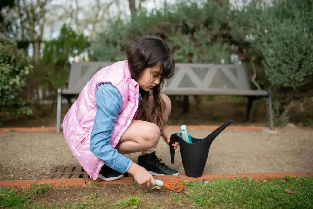 A young girl tends to a garden in spring, using a hand rake near a watering can.