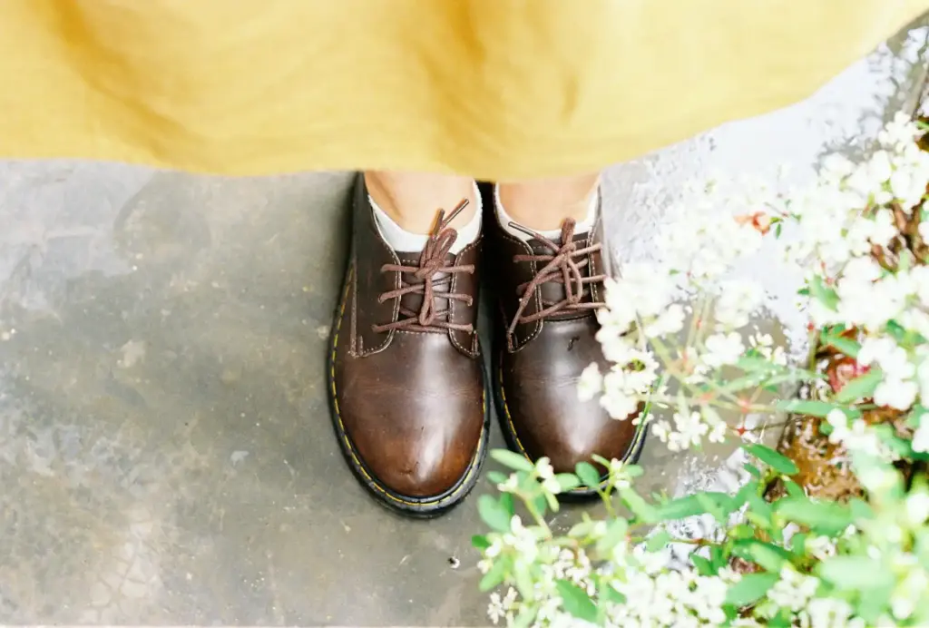 Close-up of brown leather shoes on wet ground, surrounded by small plants.