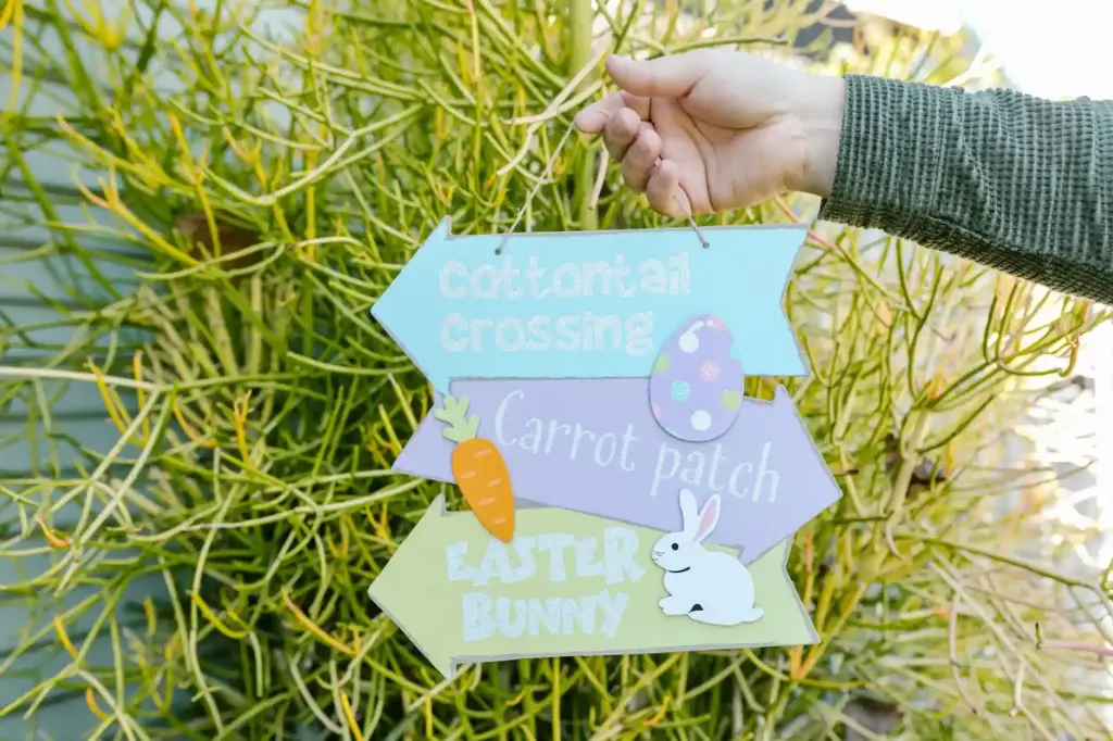 Hand holding a colorful Easter sign with bunny and carrot motifs against a plant backdrop.