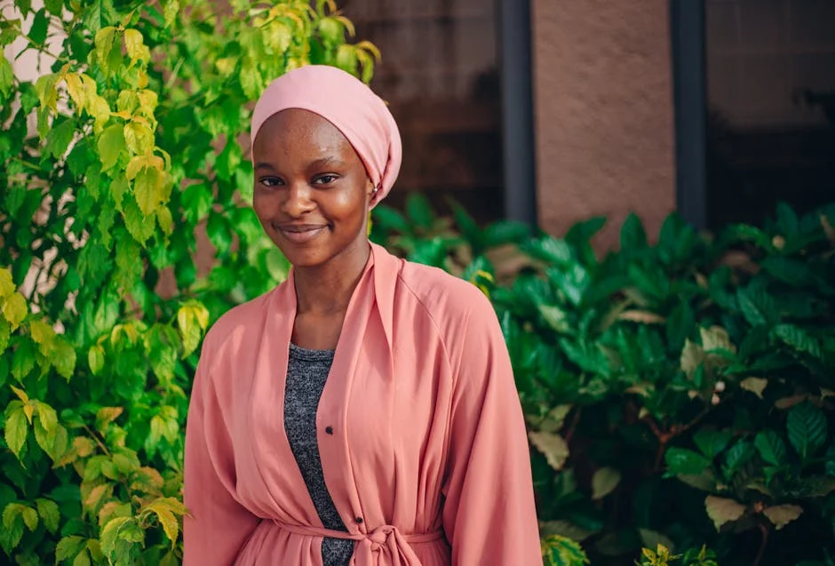 A woman in a pink hijab and robe smiles against a green leafy background outdoors.