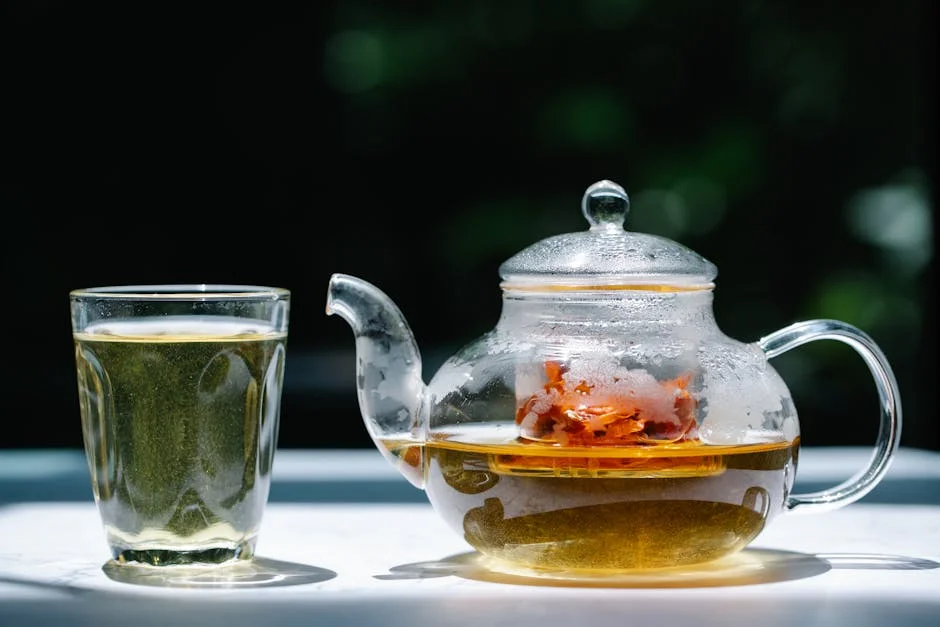 A glass teapot filled with herbal tea next to a transparent glass under sunlight. Refreshing and aromatic.