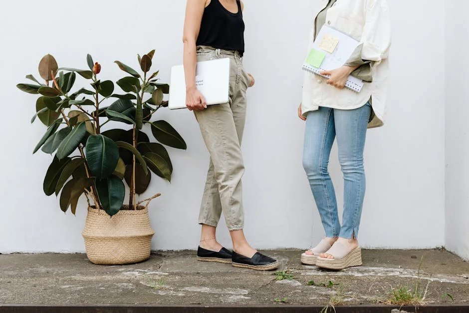 Two women standing casually, holding documents, beside a potted plant, indoors.
