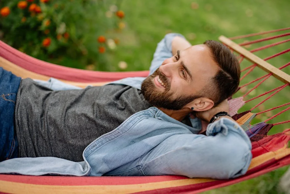 A bearded man in a denim shirt smiles while lying on a colorful hammock outdoors.