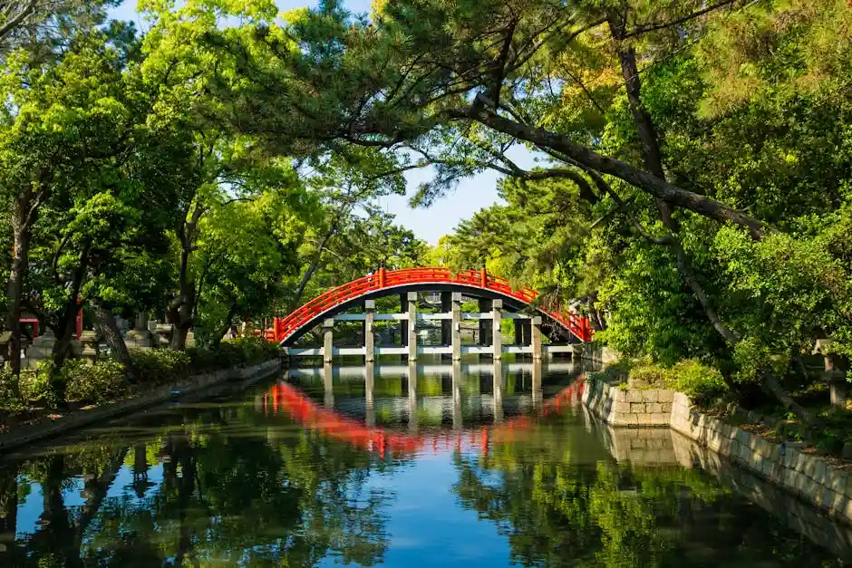 Tranquil scene of a red arched bridge in a lush, serene Japanese garden with clear reflections on a sunny day.