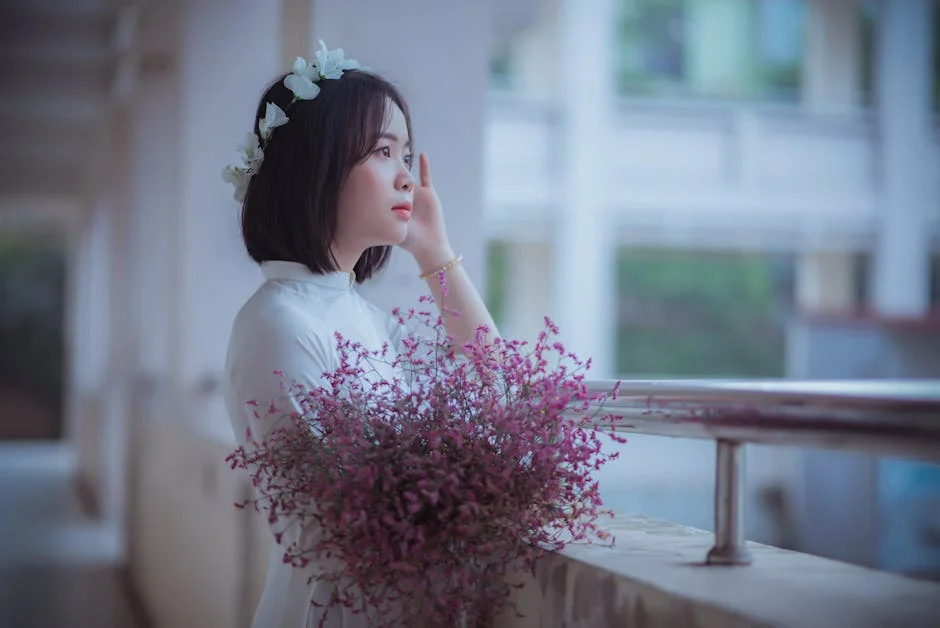 A serene Asian woman in white attire holding flowers, posing elegantly indoors.