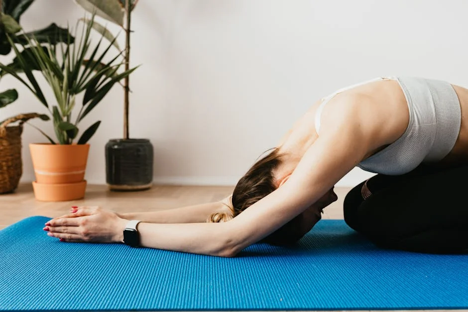 A woman in activewear practicing yoga in child pose on a blue mat indoors, promoting wellness and relaxation.