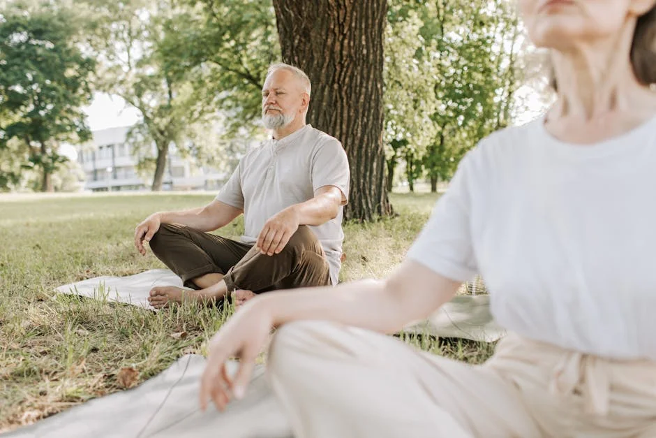 Elderly man and woman meditating outdoors on yoga mats in a peaceful park setting.