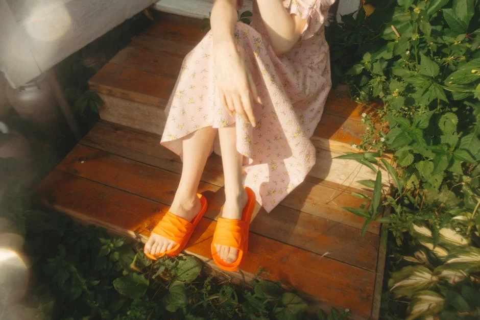 A woman in a pink floral dress with orange sandals sitting on wooden stairs surrounded by greenery.