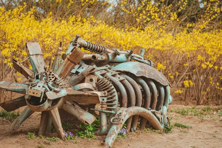 A rusty mechanical sculpture surrounded by vibrant yellow flowering shrubs in a rural setting.