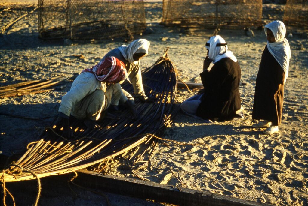 Group of people crafting traditional items in a sandy outdoor setting under sunlight.