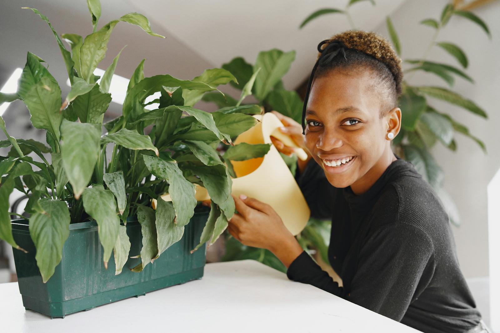 A cheerful teenager smiles while watering houseplants indoors, promoting greenery care.
