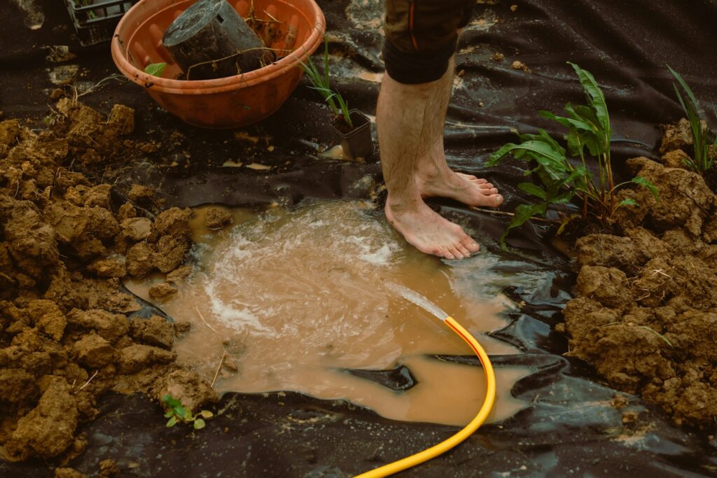 Bare feet watering muddy ground with a yellow hose