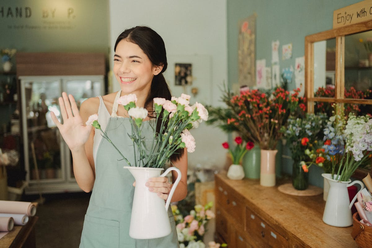 Friendly young florist waving in her shop, holding a bouquet of flowers.