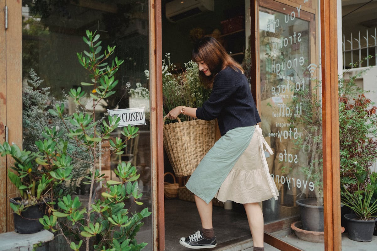 Side view of Asian female florist in casual clothes carrying basket with flowers into shop