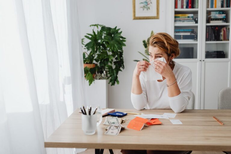 Worried woman at home desk with bills and dollar bills showing financial stress.
