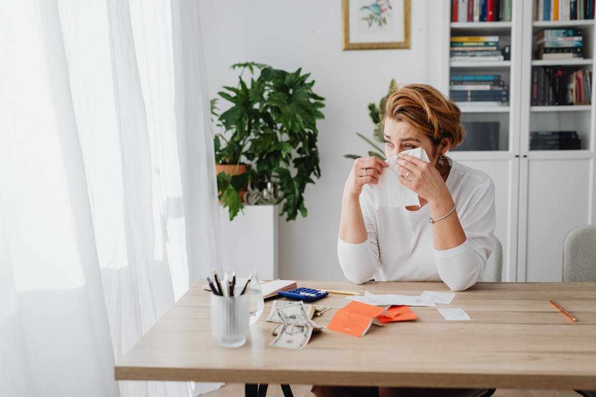Worried woman at home desk with bills and dollar bills showing financial stress.