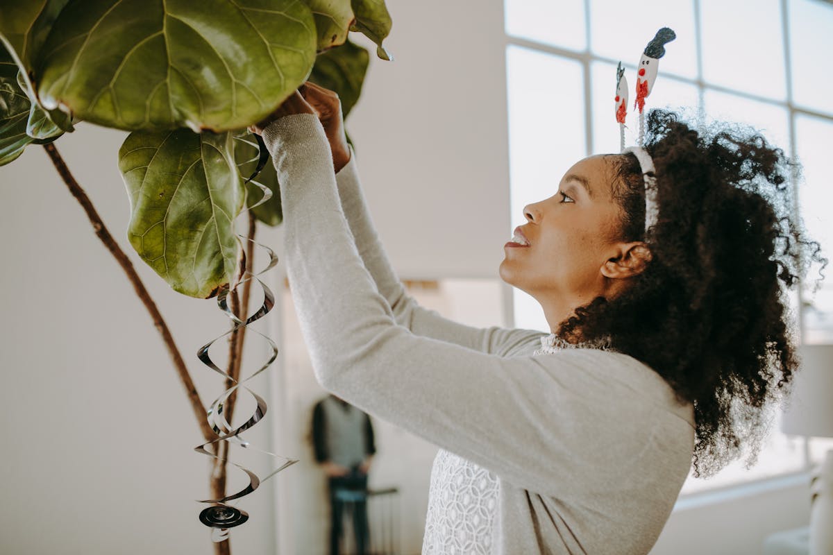 A woman decorates a large indoor plant with whimsical holiday ornaments.