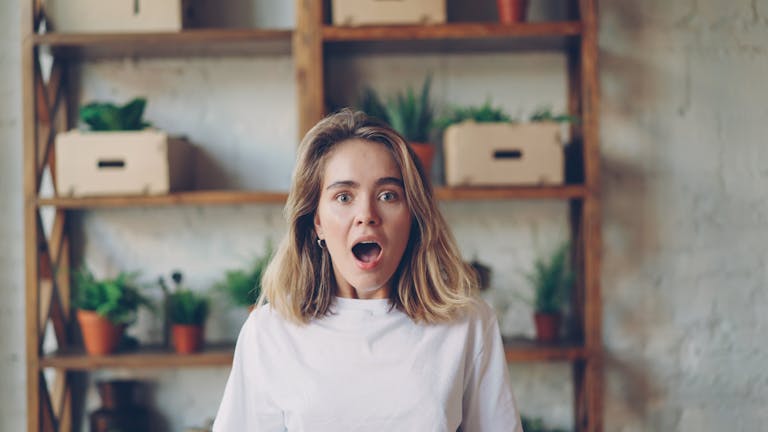 Young woman in white shirt with a surprised expression indoors, surrounded by plants.